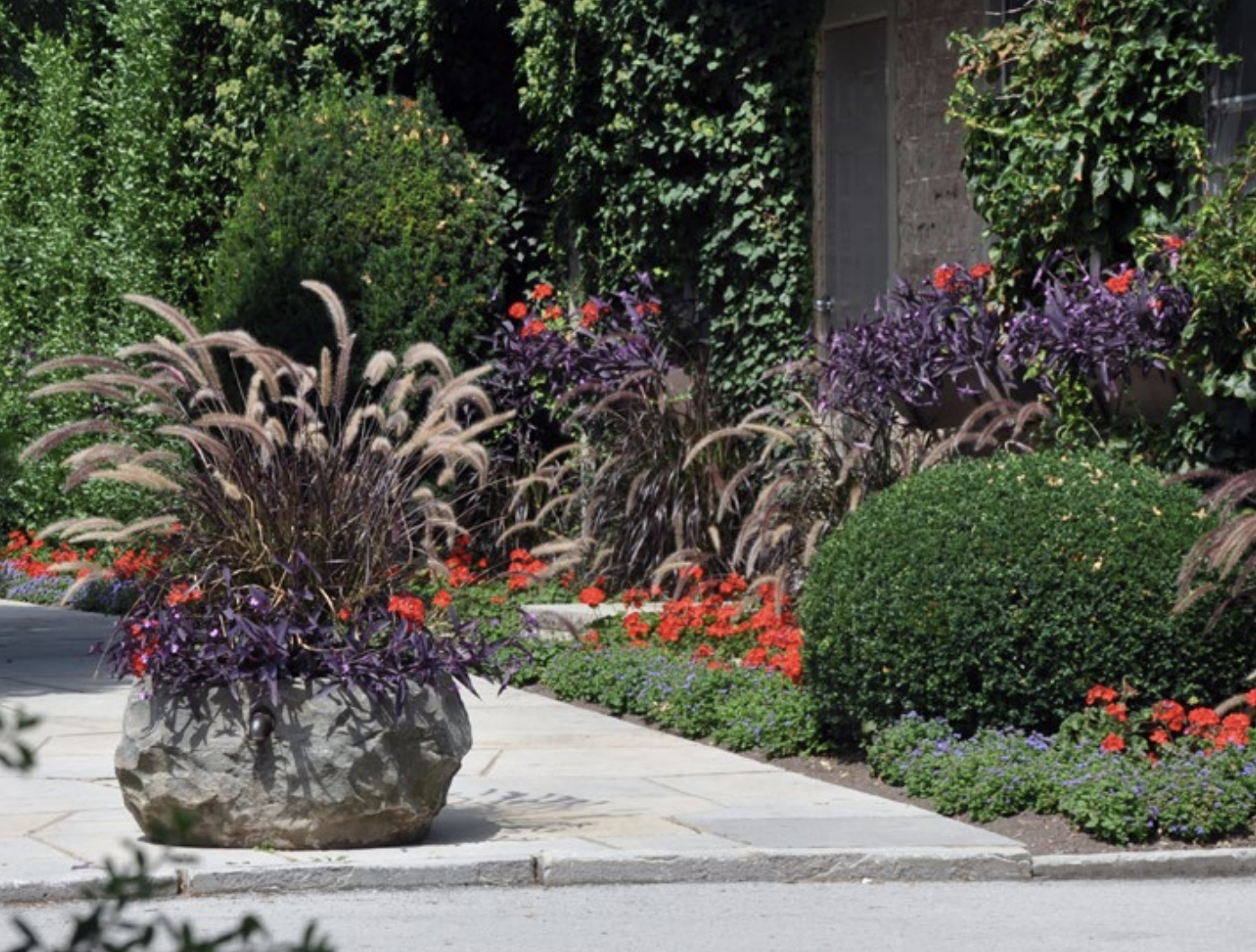 A stone garden urn filled with tall grasses and flowers in front of a garden with red flowers and bushes